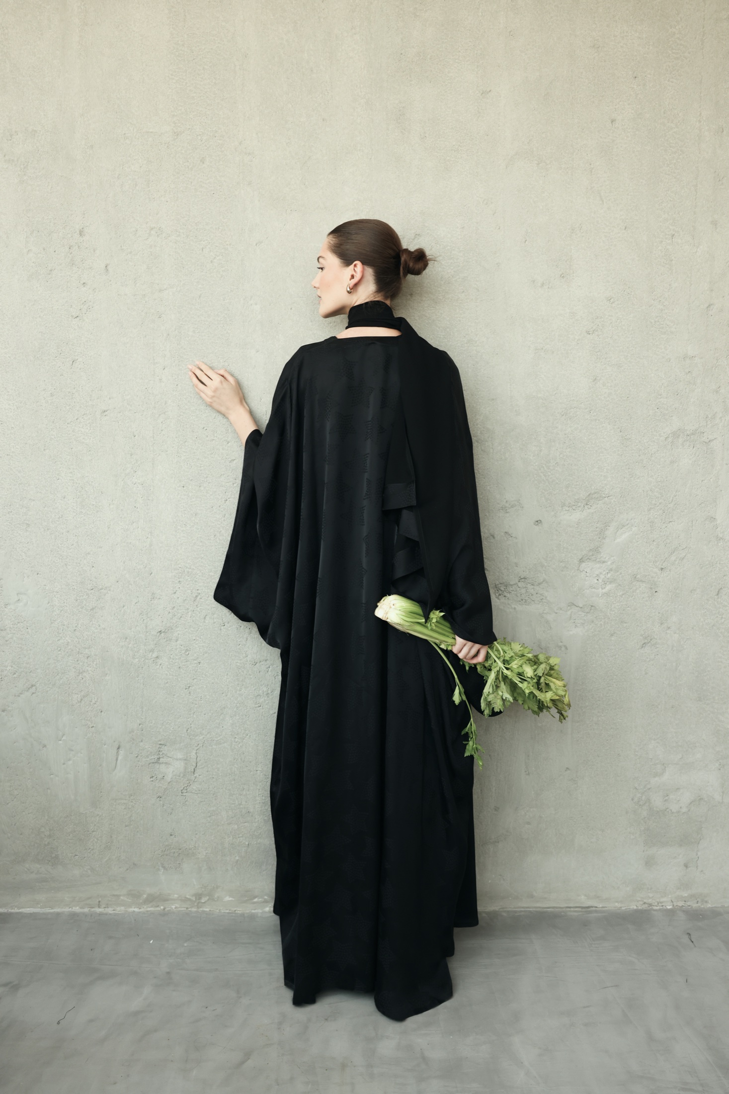 Model in a black abaya holding greens against a textured wall.